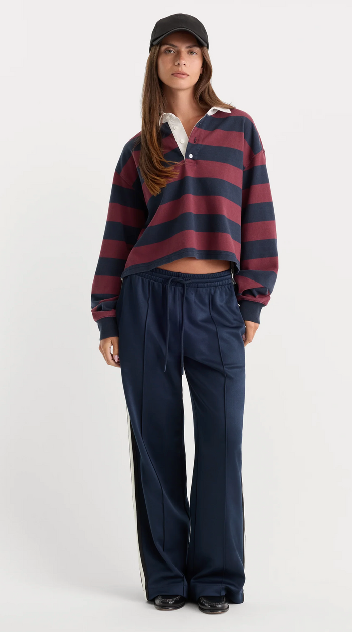 A model stands against a light gray studio background wearing a navy and burgundy striped cropped rugby shirt, navy wide-leg sweatpants, a black baseball cap, and black shoes.