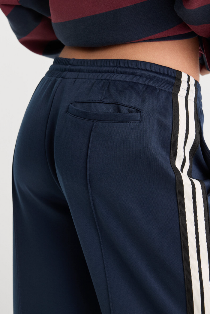 Close-up of navy blue track pants with white side stripes, shown on a model from the waist down.