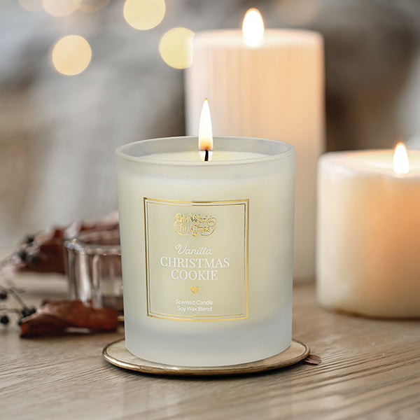 A lit vanilla Christmas cookie scented candle in a frosted glass jar sits on a table, with two larger lit candles and festive decor in the softly blurred background.