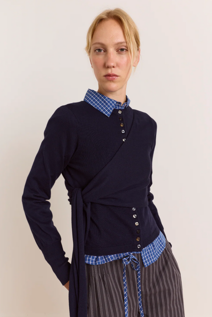 A model wearing a navy wrap-style long-sleeve top with a blue and white checkered collar and matching tie detail, paired with striped trousers, against a plain light background.