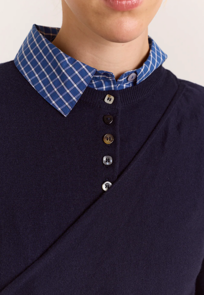 Close-up of a person wearing a navy knit sweater layered over a blue and white plaid collared shirt, with the collar and front buttons visible.