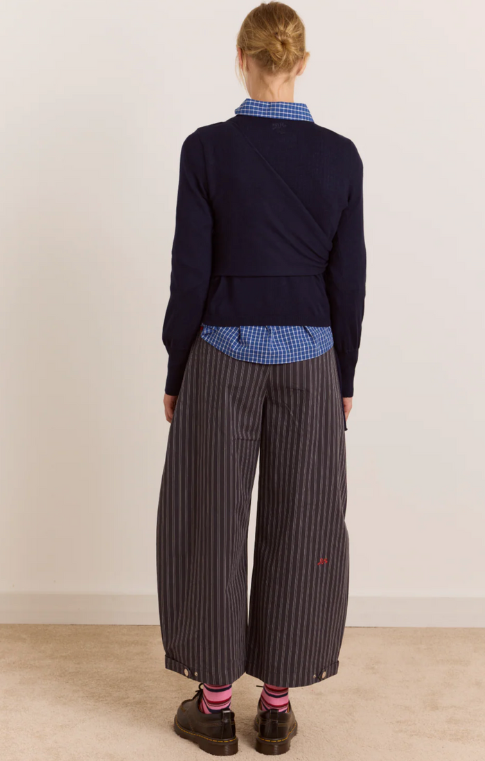 A model shown from the back wearing a navy wrap-style cardigan over a blue and white gingham shirt, paired with dark gray pinstriped wide-leg trousers, pink socks, and dark brown shoes, set against a plain off-white wall and beige floor.