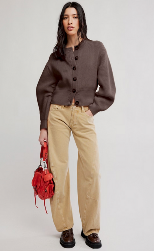 Woman wearing a coffee-colored cardigan, beige pants, and holding a red bag on a white background