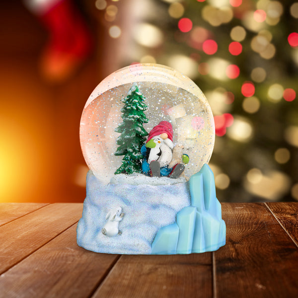 A festive snow globe featuring a penguin wearing a Santa hat sitting by a pine tree, with a white rabbit at the snowy base. The background is blurred with warm, colorful holiday lights.