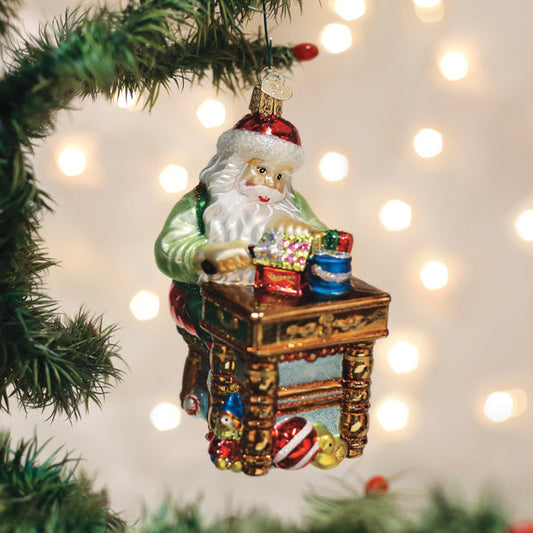 A glass ornament of Santa Claus sitting at a desk with wrapped presents, hanging on a Christmas tree branch, with blurred festive lights in the background.